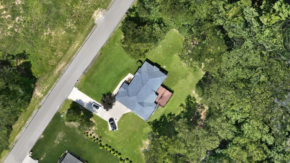 Overhead capture showing the home, driveway, road, lawn, and surrounding trees